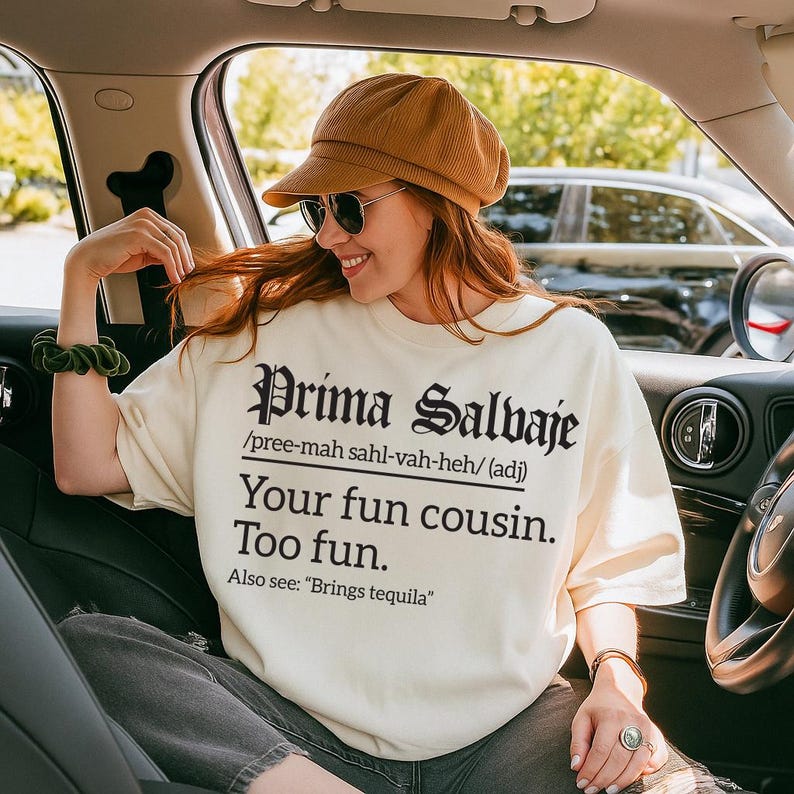 Off-white sweatshirt with black text reading “Prima Salvaje /pree-mah sahl-vah-heh/ (adj) Your fun cousin. Too fun. Also see: ‘Brings tequila,’” worn by a smiling woman sitting in a car.