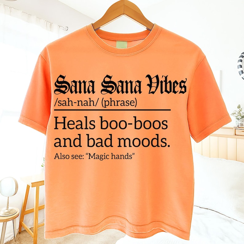 Bright orange T-shirt with bold black text reading "Sana Sana Vibes /sah-nah/ (phrase) Heals boo-boos and bad moods. Also see: 'Magic hands'" in Gothic and serif fonts, displayed on a hanger indoors.
