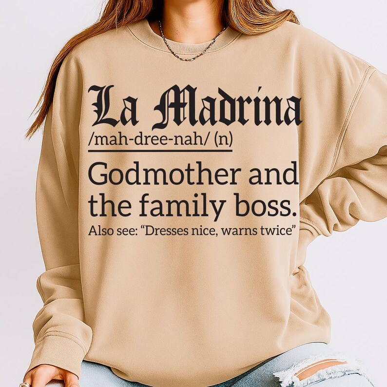 Beige sweatshirt with black text reading “La Madrina /mah-dree-nah/ (n) Godmother and the family boss. Also see: ‘Dresses nice, warns twice,’” modeled by a woman with one hand in her pocket.