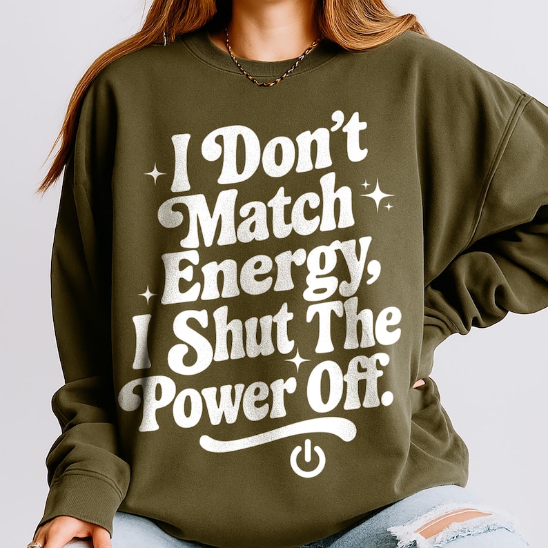 Olive green sweatshirt with bold white text saying "I Don’t Match Energy, I Shut The Power Off" in a retro groovy font, worn by a woman with wavy hair. Sassy and empowering quote shirt for women.