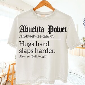 White t-shirt on a hanger with black text reading “Abuelita Power /ah-bweh-lee-tah/ (n) Hugs hard, slaps harder. Also see: ‘Built tough,’” displayed in a bright bedroom setting.