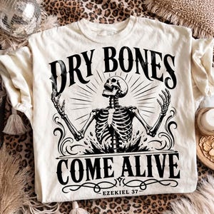 Flat lay of an off-white Christian graphic tee on a leopard print backdrop with boho disco ball decor. The center print shows an intricate black skeleton rising toward light rays with text "Dry Bones Come Alive, Ezekiel 37" and ornate flourishes. An edgy, trendy scripture graphic shirt perfect for modern faith apparel collections and edgy gifts.