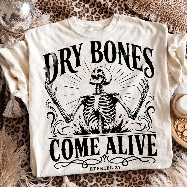 Flat lay of an off-white Christian graphic tee on a leopard print backdrop with boho disco ball decor. The center print shows an intricate black skeleton rising toward light rays with text "Dry Bones Come Alive, Ezekiel 37" and ornate flourishes. An edgy, trendy scripture graphic shirt perfect for modern faith apparel collections and edgy gifts.