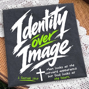 Flat lay of a neatly folded dark charcoal gray t-shirt resting on a woven basket with white lace detailing. The shirt showcases a bold white and neon green brush typography design reading "Identity Over Image" with "1 Samuel 16:7" and a simple cross illustration. A unique scripture graphic tee emphasizing inner faith and spiritual value over outward appearance.