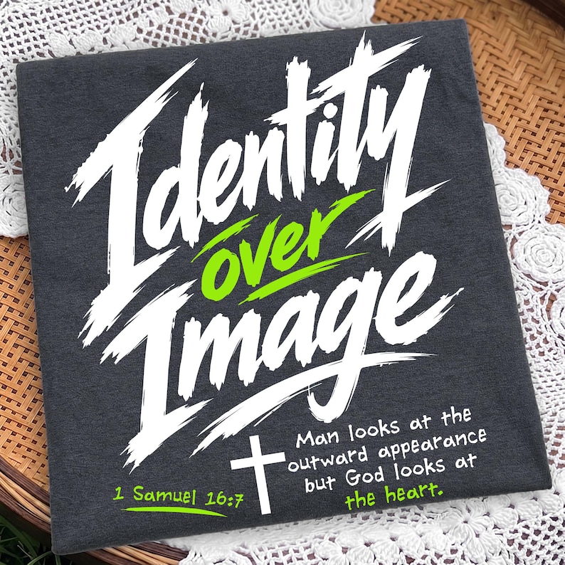 Flat lay of a neatly folded dark charcoal gray t-shirt resting on a woven basket with white lace detailing. The shirt showcases a bold white and neon green brush typography design reading "Identity Over Image" with "1 Samuel 16:7" and a simple cross illustration. A unique scripture graphic tee emphasizing inner faith and spiritual value over outward appearance.