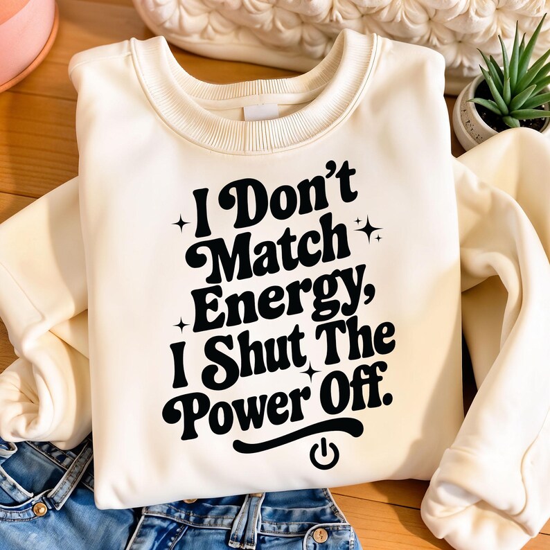 Neutral sweatshirt with bold black “I Don’t Match Energy, I Shut the Power Off” quote in playful retro font, folded neatly next to denim – ideal for sublimation or DTF design preview.