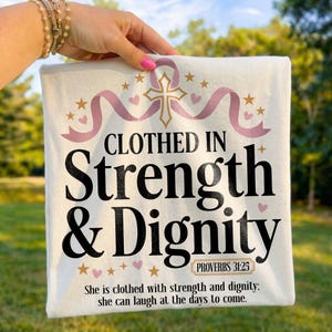 Könnte beinhalten: Ein weißes T-Shirt mit einer Grafik aus einem goldenen Kreuz, rosa Bändern und kleinen goldenen Sternen. Der Text lautet: CLOTHED IN Strength & Dignity, PROVERBS 31:25, und She is clothed with strength and dignity: she can laugh at the days to come.
