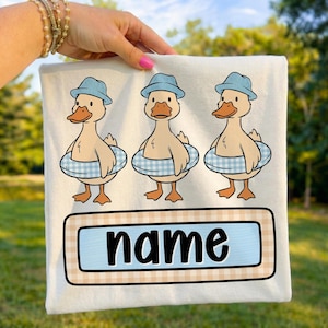 May include: A white t-shirt featuring three cartoon ducks wearing blue hats and inflatable rings. The ducks have orange beaks and feet. Below the ducks is a rectangular design with the word "name" in black letters.