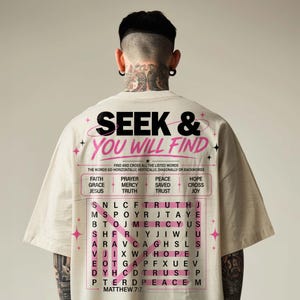 Back view of an oversized cream T-shirt on a male model, showcasing a large Christian “Seek & You Will Find” word search graphic in black and pink. The design features Matthew 7:7, faith words like prayer, grace, mercy, hope, cross, and Jesus, plus modern star accents for a bold streetwear-inspired Christian shirt mockup. Clean neutral studio background.