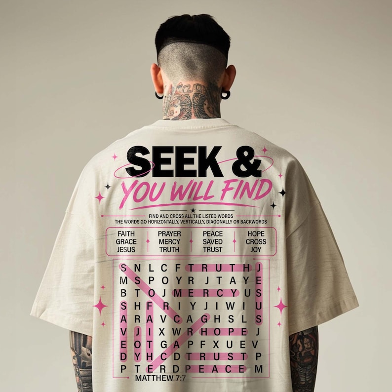 Back view of an oversized cream T-shirt on a male model, showcasing a large Christian “Seek & You Will Find” word search graphic in black and pink. The design features Matthew 7:7, faith words like prayer, grace, mercy, hope, cross, and Jesus, plus modern star accents for a bold streetwear-inspired Christian shirt mockup. Clean neutral studio background.