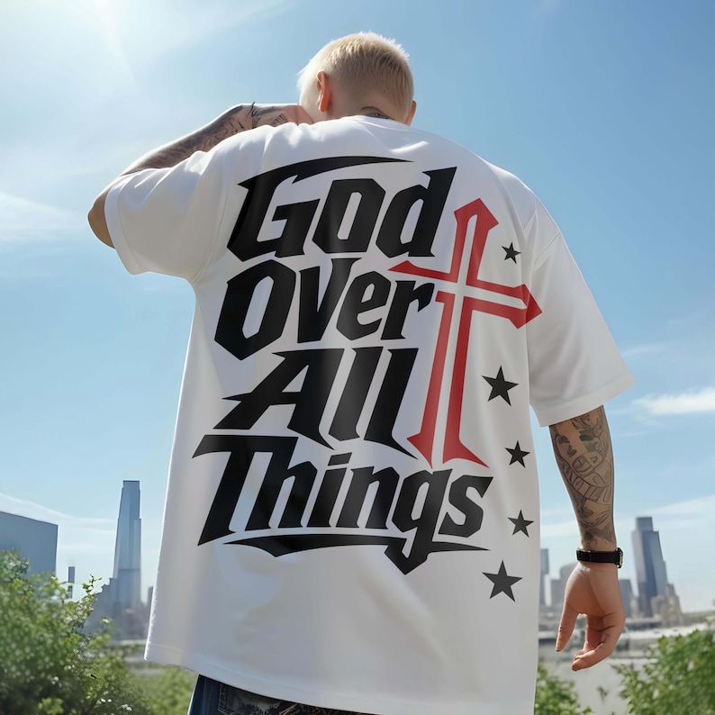 Back view of a model wearing an oversized white streetwear t-shirt outdoors under a sunny sky with city buildings. The back displays sharp black lettering reading "God Over All Things" alongside a bold red cross graphic and black star accents. A trendy, urban Christian apparel option designed for an eye-catching faith-filled streetwear style.