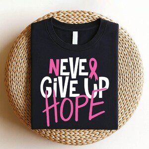 May include: Black t-shirt with the words "NEVER GIVE UP HOPE" in white and pink lettering. A pink ribbon is incorporated into the design. The shirt is displayed on a woven, circular surface.