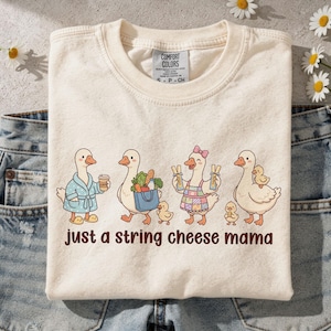 May include: A cream-coloured t-shirt with a playful design of four geese, each with distinct accessories, and the text "just a string cheese mama." The geese are shown with a coffee cup, groceries, and a baby chick.