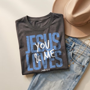 Charcoal dark gray folded t-shirt mockup featuring a distressed blue and white “Jesus Loves You & Me” Christian typography graphic with John 15:13 scripture. The shirt is styled with light denim jeans, layered gold necklaces, and a beige hat in a neutral lifestyle flat lay scene. This mockup highlights a bold faith-based graphic tee ideal for Christian apparel listings and inspirational clothing designs.