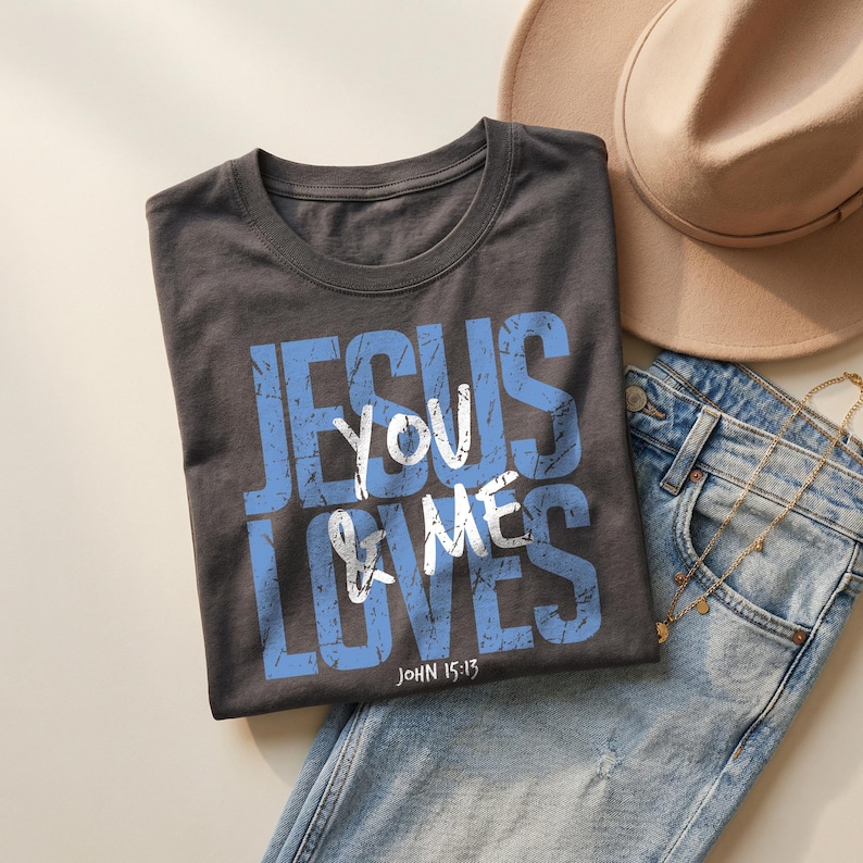 Charcoal dark gray folded t-shirt mockup featuring a distressed blue and white “Jesus Loves You & Me” Christian typography graphic with John 15:13 scripture. The shirt is styled with light denim jeans, layered gold necklaces, and a beige hat in a neutral lifestyle flat lay scene. This mockup highlights a bold faith-based graphic tee ideal for Christian apparel listings and inspirational clothing designs.
