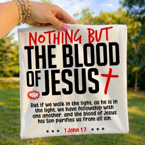 Folded white t-shirt held outdoors by a hand with pink nails against a blurred green background. The shirt displays bold red and black text reading "Nothing But The Blood of Jesus" alongside a red cross, crown of thorns, and the verse "1 John 1:7". A crisp, vibrant faith-based graphic tee highlighting a central Christian gospel message for everyday wear.