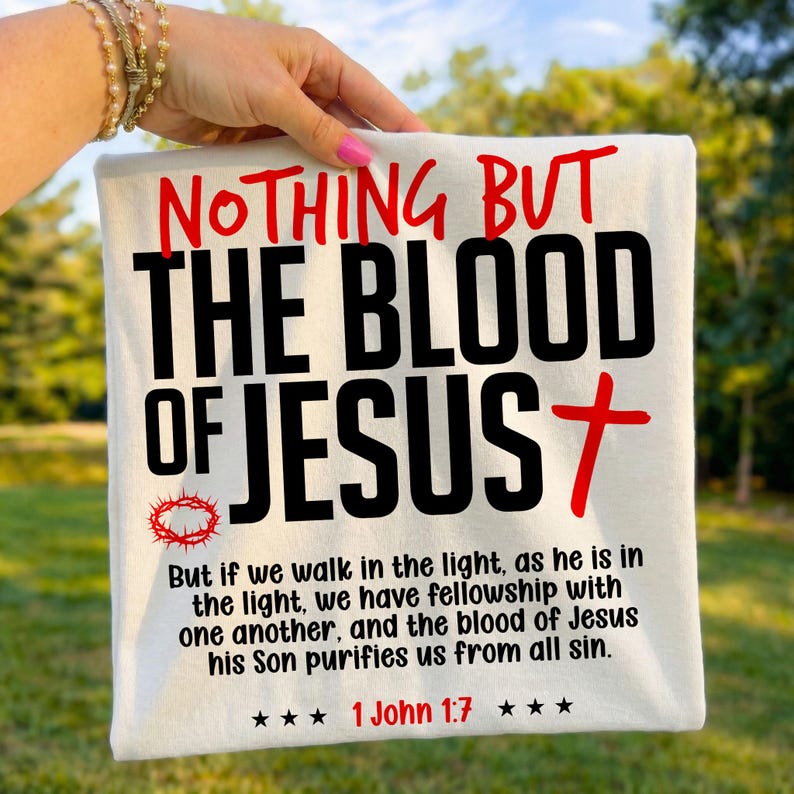 Folded white t-shirt held outdoors by a hand with pink nails against a blurred green background. The shirt displays bold red and black text reading "Nothing But The Blood of Jesus" alongside a red cross, crown of thorns, and the verse "1 John 1:7". A crisp, vibrant faith-based graphic tee highlighting a central Christian gospel message for everyday wear.