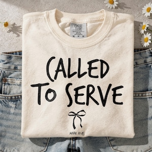 May include: Cream-colored t-shirt with the words "CALLED TO SERVE" in black script, a bow graphic, and the Bible verse Mark 10:45. The shirt is folded on denim jeans, with daisies scattered around.