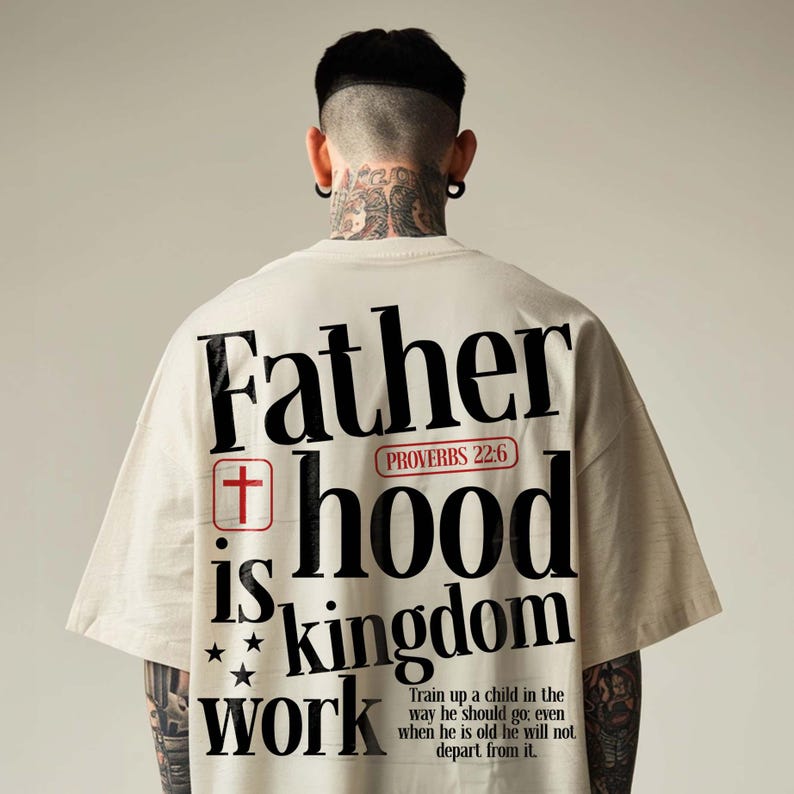 Back view of a tattooed man with an undercut hairstyle wearing an oversized cream t-shirt. The back of the shirt features a large, bold typographic Christian graphic that reads "Father hood is kingdom work" in black block letters. The design includes a small red square with a cross, a red Proverbs 22:6 badge, three black stars, and the full verse quote printed at the bottom against a neutral studio background.