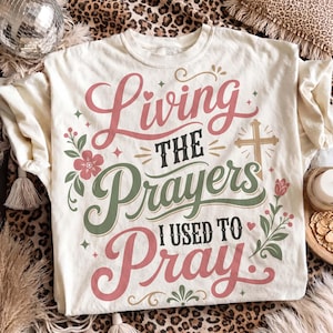 Flat lay presentation of an off-white graphic tee styled on a leopard print backdrop with boho accents. The shirt displays beautiful pink and sage green script lettering reading "Living the Prayers I Used to Pray" adorned with a cross and floral accents. A sweet, aesthetic Christian graphic t-shirt ideal for women celebrating answered prayers.