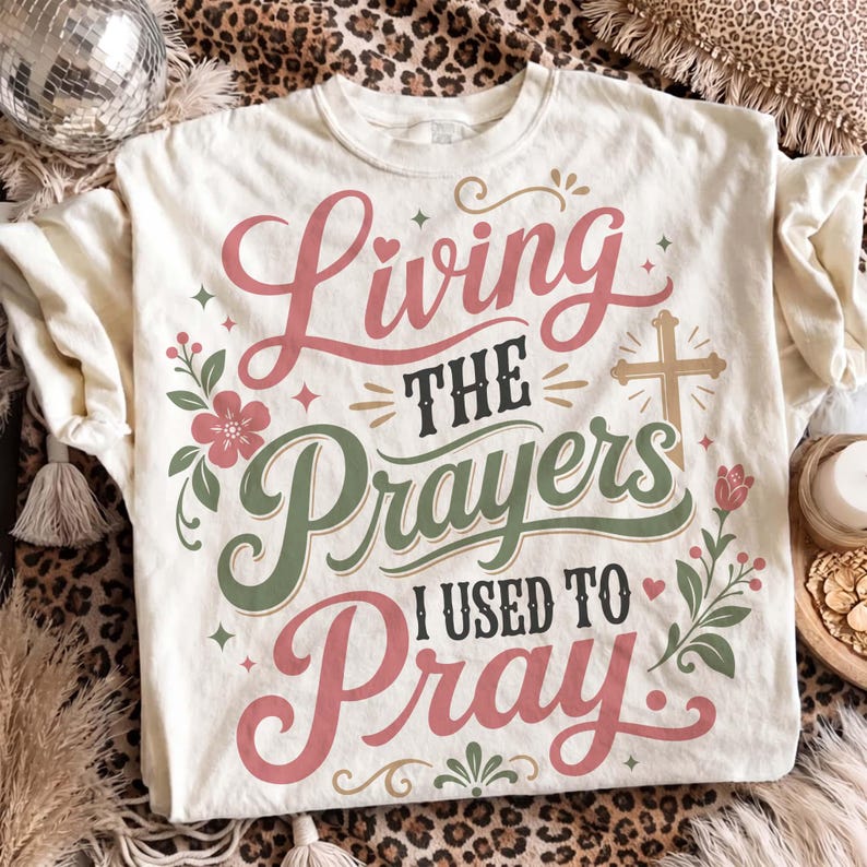 Flat lay presentation of an off-white graphic tee styled on a leopard print backdrop with boho accents. The shirt displays beautiful pink and sage green script lettering reading "Living the Prayers I Used to Pray" adorned with a cross and floral accents. A sweet, aesthetic Christian graphic t-shirt ideal for women celebrating answered prayers.