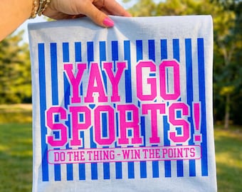 Op de afbeelding: Een lichtblauw t-shirt met verticale blauwe strepen en de woorden "YAY GO SPORTS!" in felroze. Onder de hoofdtekst staat de zin "DO THE THING - WIN THE POINTS" in roze. Het shirt wordt voor een groene achtergrond gehouden.