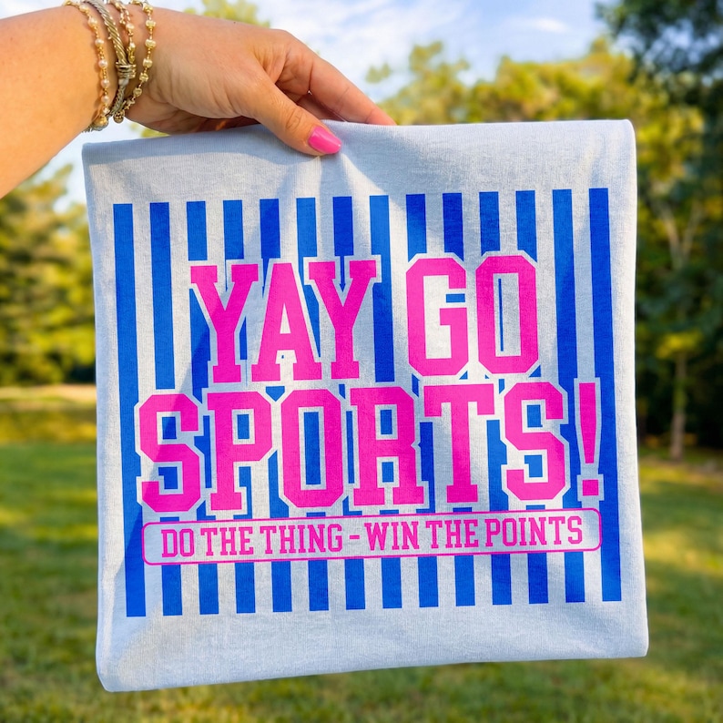 Close-up lifestyle mockup of a folded white t-shirt held outdoors against a soft green background, featuring a bold “Yay Go Sports!” graphic in hot pink varsity lettering with vertical royal blue stripes and the phrase “Do The Thing – Win The Points” underneath, creating a vibrant game day sports fan shirt design with bright contrast and cheerful energy.