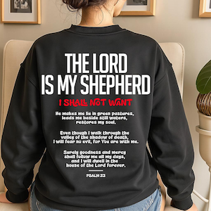 Back view of a woman wearing a black Christian crewneck sweatshirt in a cozy indoor setting. The back print features bold white and red typography reading "The Lord Is My Shepherd I Shall Not Want" followed by the full text of Psalm 23 scripture. A comfortable, inspirational faith jumper offering a powerful biblical reminder for daily wear.