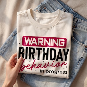 以下が含まれることがあります： 「WARNING BIRTHDAY behavior in progress」の文字が黒とピンクでプリントされた、クリーム色のTシャツ。Tシャツは折りたたまれ、青いジーンズの上に置かれています。