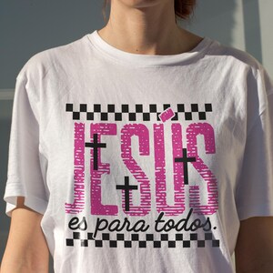 White oversized women’s t-shirt worn outdoors in golden sunlight, showcasing vibrant pink “JESÚS” typography with black cross details and checkerboard borders, paired with “es para todos.” beneath. This Spanish Christian statement shirt combines modern faith fashion with casual everyday styling, ideal for worship events and inspirational wear.