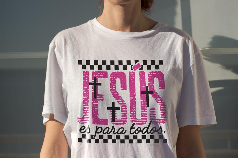 White oversized women’s t-shirt worn outdoors in golden sunlight, showcasing vibrant pink “JESÚS” typography with black cross details and checkerboard borders, paired with “es para todos.” beneath. This Spanish Christian statement shirt combines modern faith fashion with casual everyday styling, ideal for worship events and inspirational wear.