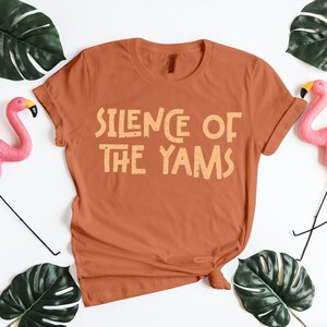 Rust orange fall T-shirt printed with “Silence of the Yams” in beige distressed letters. Funny Thanksgiving shirt mixing humor with holiday puns. Perfect for Friendsgiving, turkey day cooking, or matching family outfits. Great gift for food lovers and Thanksgiving PNG design fans who adore playful seasonal quotes on cozy autumn apparel.