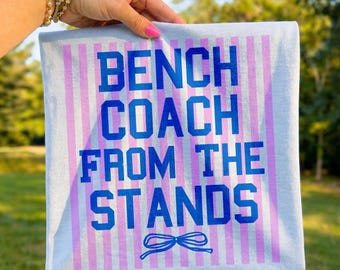 Op de afbeelding: Een lichtblauwe rechthoekige handdoek met verticale roze strepen en de tekst "BENCH COACH FROM THE STANDS" in blauw. Een blauwe strik-afbeelding staat onderaan. De handdoek wordt door een persoon omhoog gehouden.