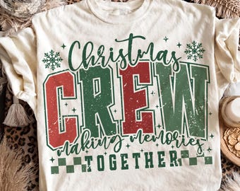 Op de afbeelding: Een crèmekleurig T-shirt met de tekst Christmas CREW making memories TOGETHER in rode en groene letters met een verweerde uitstraling. Het ontwerp bevat sneeuwvlokpictogrammen, kleine steraccenten en een dambordpatroon onder het woord TOGETHER.