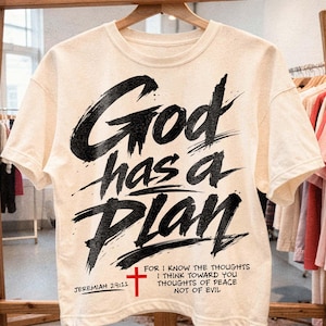 Cream oversized T-shirt hanging on a wooden hanger in a boutique shop setting with clothing racks softly blurred in the background. The front features a bold black brush-style Christian design reading “God has a Plan,” finished with a red cross, Jeremiah 29:11 reference, and a compact scripture quote along the lower portion for a clean faith-based streetwear look.