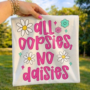 May include: Cream-colored t-shirt with the phrase "all oopsies, no daisies" in bright pink lettering. The design includes daisy and flower graphics in white, yellow, and teal. The shirt is held up by a person.
