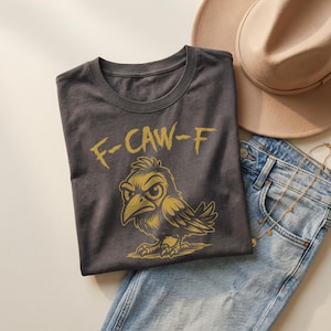 Flat lay of charcoal gray t-shirt folded on denim jeans beside beige hat and layered gold jewelry. Metallic gold “F-CAW-F” text and crow illustration create a striking contrast against dark fabric. Soft lighting and neutral styling give this edgy funny bird shirt a modern boutique aesthetic.