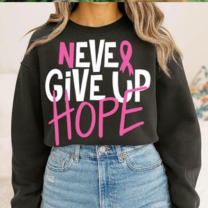 May include: Black sweatshirt with the words "NEVER GIVE UP HOPE" in white and pink lettering. The design includes a pink ribbon, a symbol of breast cancer awareness. The sweatshirt is paired with blue denim jeans.