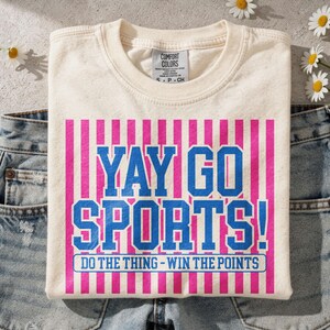 Flat lay mockup of a cream Comfort Colors t-shirt folded over light wash denim jeans with small white daisies nearby, showcasing a distressed “Yay Go Sports!” design in blue and pink varsity typography with vertical stripe background and “Do The Thing – Win The Points” slogan, styled for a trendy casual sports fan aesthetic.