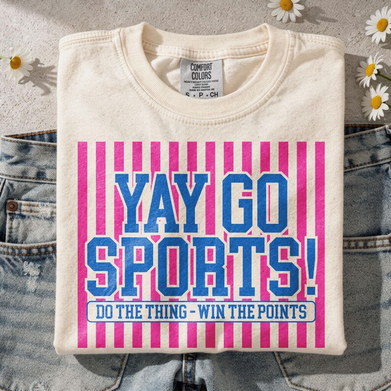 Flat lay mockup of a cream Comfort Colors t-shirt folded over light wash denim jeans with small white daisies nearby, showcasing a distressed “Yay Go Sports!” design in blue and pink varsity typography with vertical stripe background and “Do The Thing – Win The Points” slogan, styled for a trendy casual sports fan aesthetic.