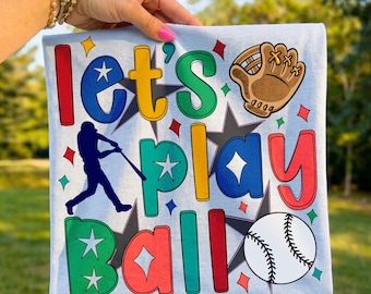 Op de afbeelding: Lichtblauw honkbal-thema T-shirt met de woorden "Let's Play Ball" in kleurrijke letters. Voorzien van een honkbalhandschoen, honkbal en een silhouet van een slagman. Het ontwerp bevat sterren en sterren.
