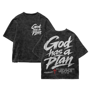 Black acid-wash oversized T-shirt mockup showing both front and back views on a plain white background. The front has a small chest print reading “God has a Plan,” while the back displays the same phrase in large white brush lettering with a red cross, Jeremiah 29:11 reference, and scripture text, creating a modern Christian streetwear design for men.