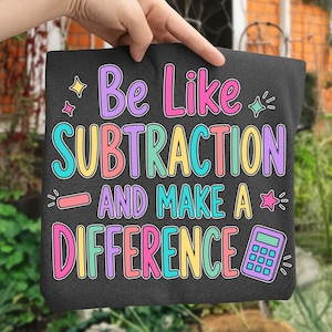 May include: A dark grey t-shirt featuring the colourful text "Be Like SUBTRACTION AND MAKE A DIFFERENCE" in pink, purple, yellow, and teal. The design includes a small minus sign, a calculator icon, and several star-shaped accents.