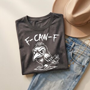 Flat lay of charcoal gray t-shirt folded on denim jeans beside beige hat and layered gold jewelry. White “F-CAW-F” text and crow illustration create a striking contrast against dark fabric. Soft lighting and neutral styling give this edgy funny bird shirt a modern boutique aesthetic.
