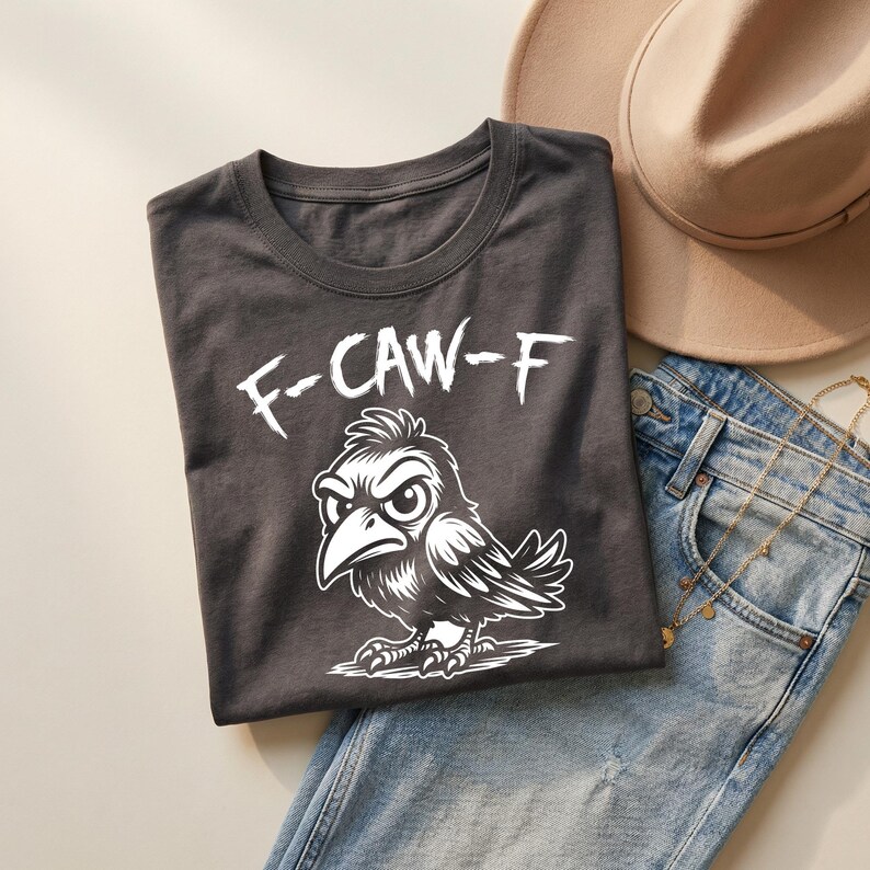 Flat lay of charcoal gray t-shirt folded on denim jeans beside beige hat and layered gold jewelry. White “F-CAW-F” text and crow illustration create a striking contrast against dark fabric. Soft lighting and neutral styling give this edgy funny bird shirt a modern boutique aesthetic.