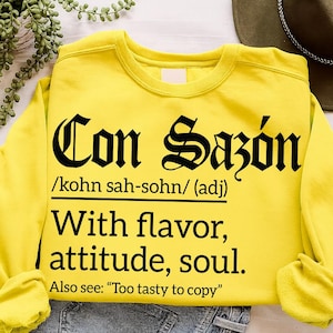 Bright yellow sweatshirt with bold black text reading "Con Sazón /kohn sah-sohn/ (adj) With flavor, attitude, soul. Also see: 'Too tasty to copy'" in Gothic and serif fonts.