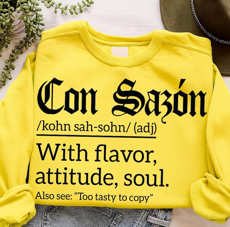 Bright yellow sweatshirt with bold black text reading "Con Sazón /kohn sah-sohn/ (adj) With flavor, attitude, soul. Also see: 'Too tasty to copy'" in Gothic and serif fonts.