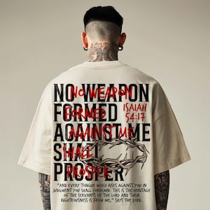 Back view of a model in an oversized cream t-shirt with neck tattoos against a neutral studio backdrop. The back print features bold black block text layered with red handwriting: "No Weapon Formed Against Me Shall Prosper, Isaiah 54:17" with a crown of thorns illustration. A bold urban Christian streetwear tee delivering an empowering faith statement.