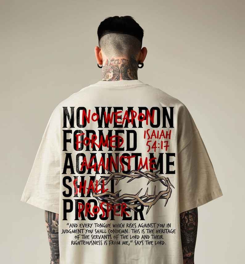 Back view of a model in an oversized cream t-shirt with neck tattoos against a neutral studio backdrop. The back print features bold black block text layered with red handwriting: "No Weapon Formed Against Me Shall Prosper, Isaiah 54:17" with a crown of thorns illustration. A bold urban Christian streetwear tee delivering an empowering faith statement.
