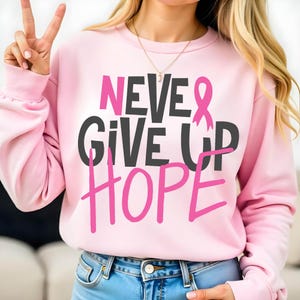 May include: A light pink sweatshirt with the words "NEVER GIVE UP HOPE" in pink and dark gray lettering. A pink ribbon graphic is included. The sweatshirt is a long-sleeved crew neck.